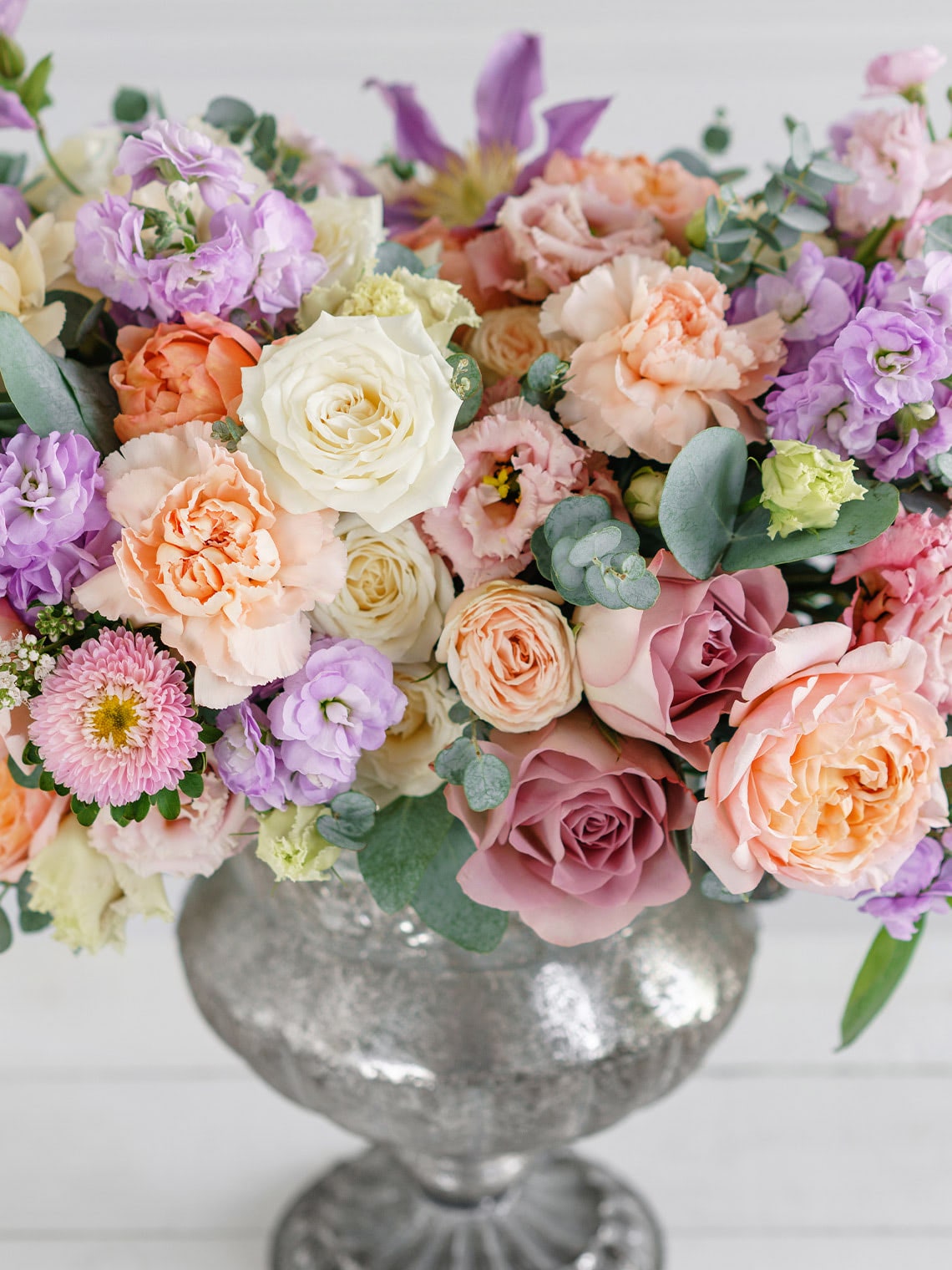 Pastel floral arrangement with roses, carnations, stock, lisianthus, eucalyptus, and seasonal blooms in a silver pedestal vase
